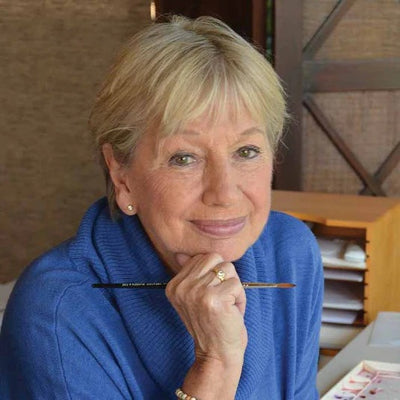 Headshot of artist Marjolein Bastin, smiling gently while holding a paintbrush, wearing a blue sweater in a cozy, studio-like setting.