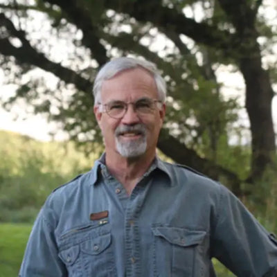 Headshot of artist Michael Sieve wearing a denim shirt and glasses, standing outdoors with trees and greenery in the background.