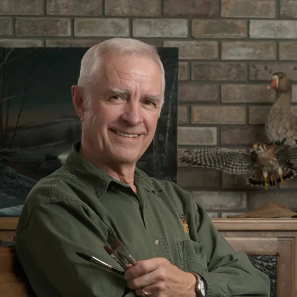 Headshot of artist Terry Redlin, smiling and holding paintbrushes while seated in front of a brick wall with wildlife-themed decor.